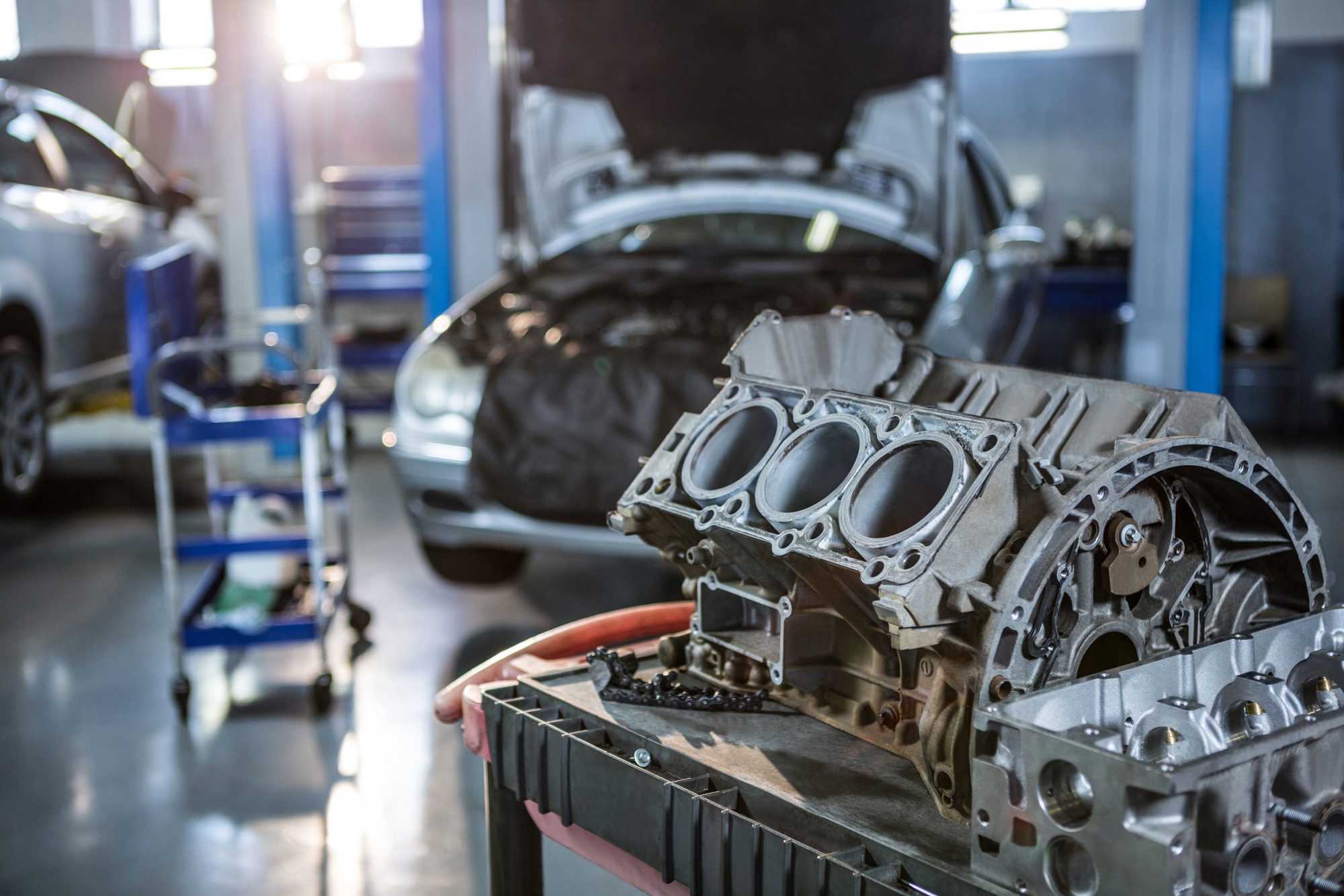 Motorblock auf der Werkbank, im Hintergrund ein Auto mit geöffneter Motorhaube in der Werkstatt
